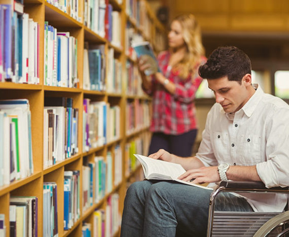 Chico en silla de ruedas leyendo un libro en la biblioteca frente a la estantería