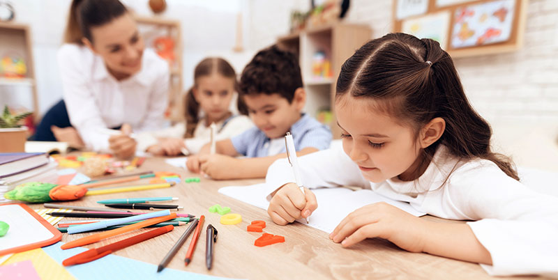 Material escolar en primer plano de una niña a la que se ve trabaja aplicadamente. Diversos lápices y bolígrafos estan delante de ella.